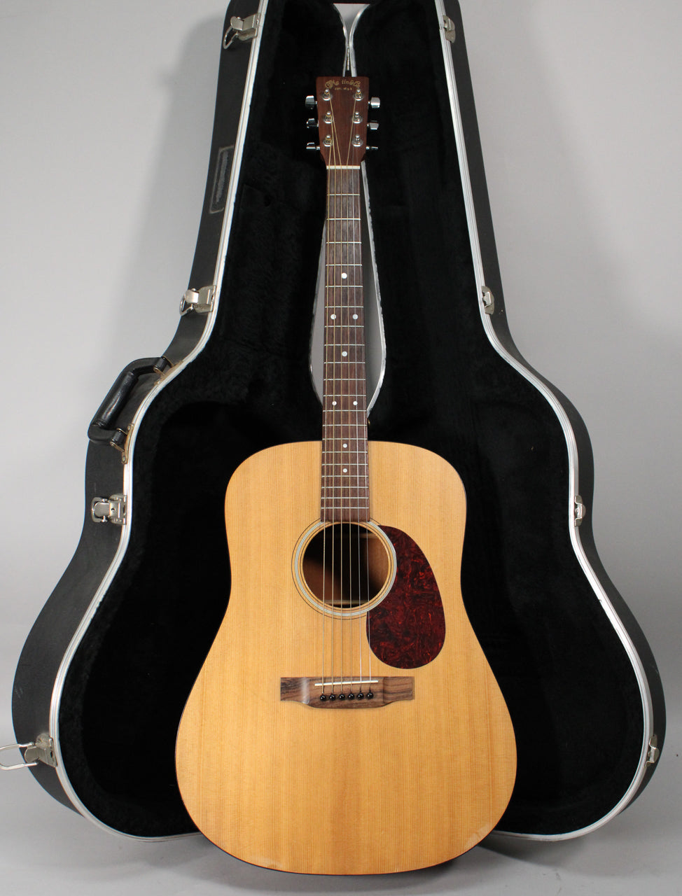 Full front view of a light wood dreadnought acoustic guitar with a dark tortoiseshell pickguard, standing in an open black hard case.