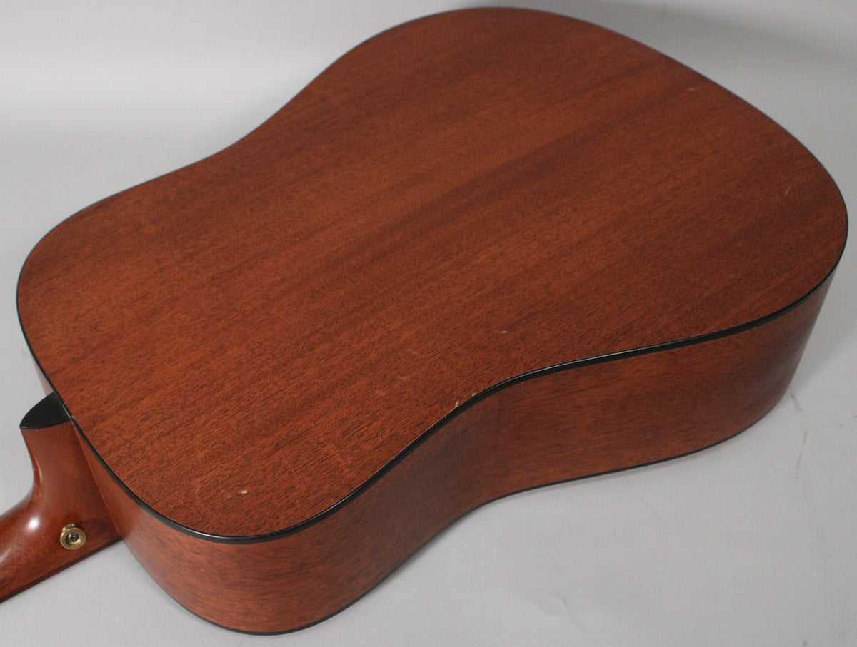 The acoustic guitar's dark brown satin mahogany back and sides are visible, with a few small dings and a strap button on the heel.