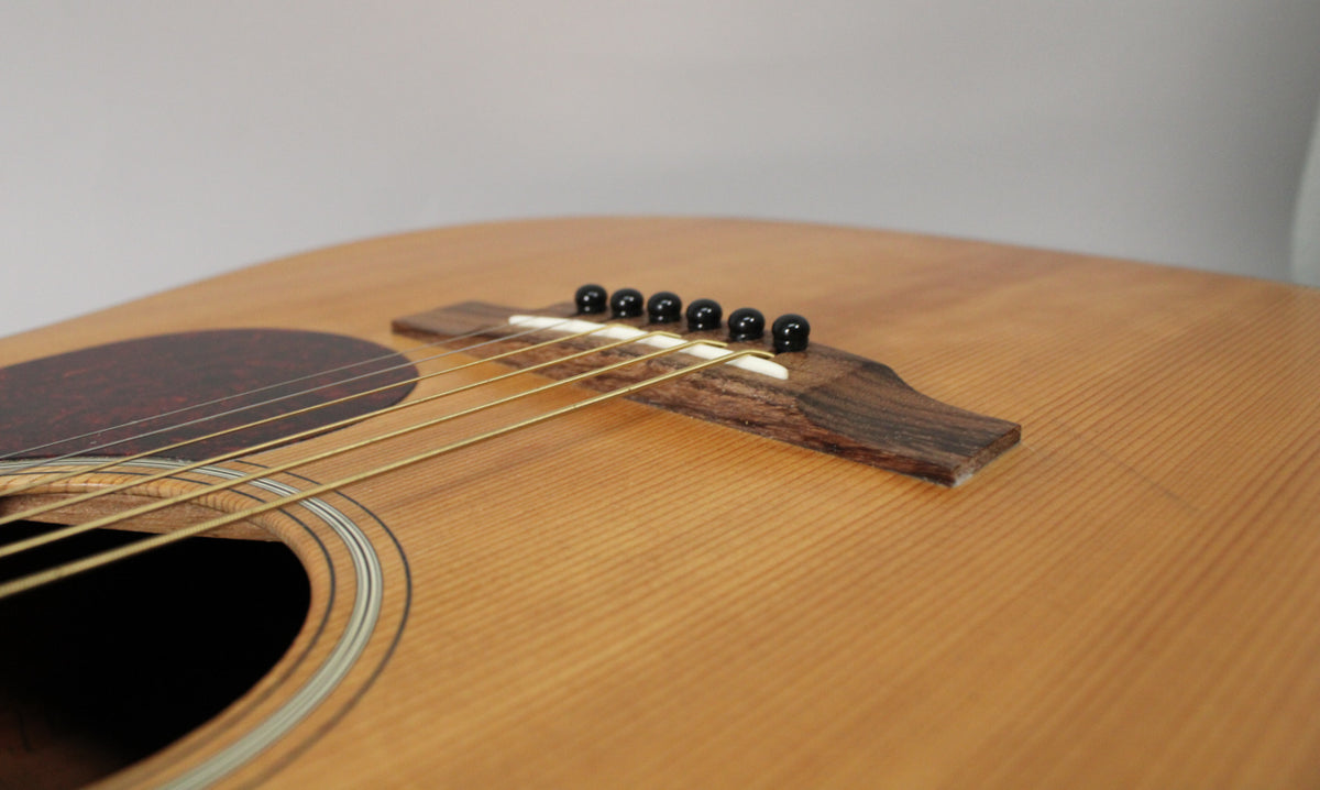 Close-up of the light brown satin top, showing the dark wood bridge with black pins, gold strings, and a round soundhole.