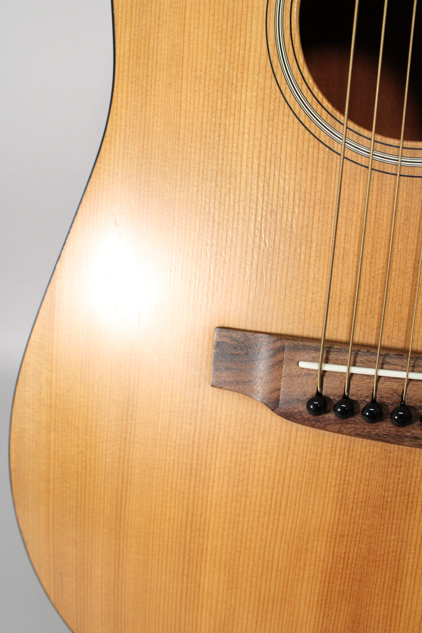 Close-up of the guitar's light-colored satin wood body, showing the dark wood bridge, white saddle, black bridge pins, and soundhole rosette.