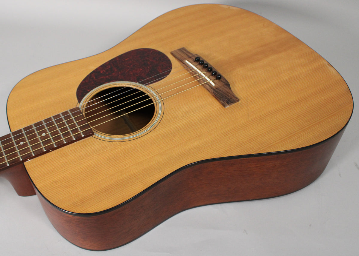 Angled view of a light wood dreadnought acoustic guitar, showing the body, dark pickguard, soundhole, bridge, and lower fretboard.