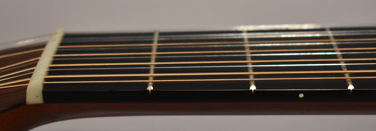 Close-up of the guitar's dark fretboard and light nut, showing 12 bronze strings and small white fret markers.