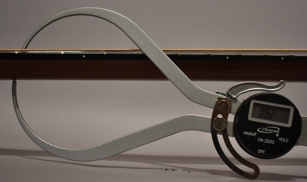 A digital caliper measures the guitar's dark brown neck, showing a reading of 0.82 inches, with the fretboard and frets visible.