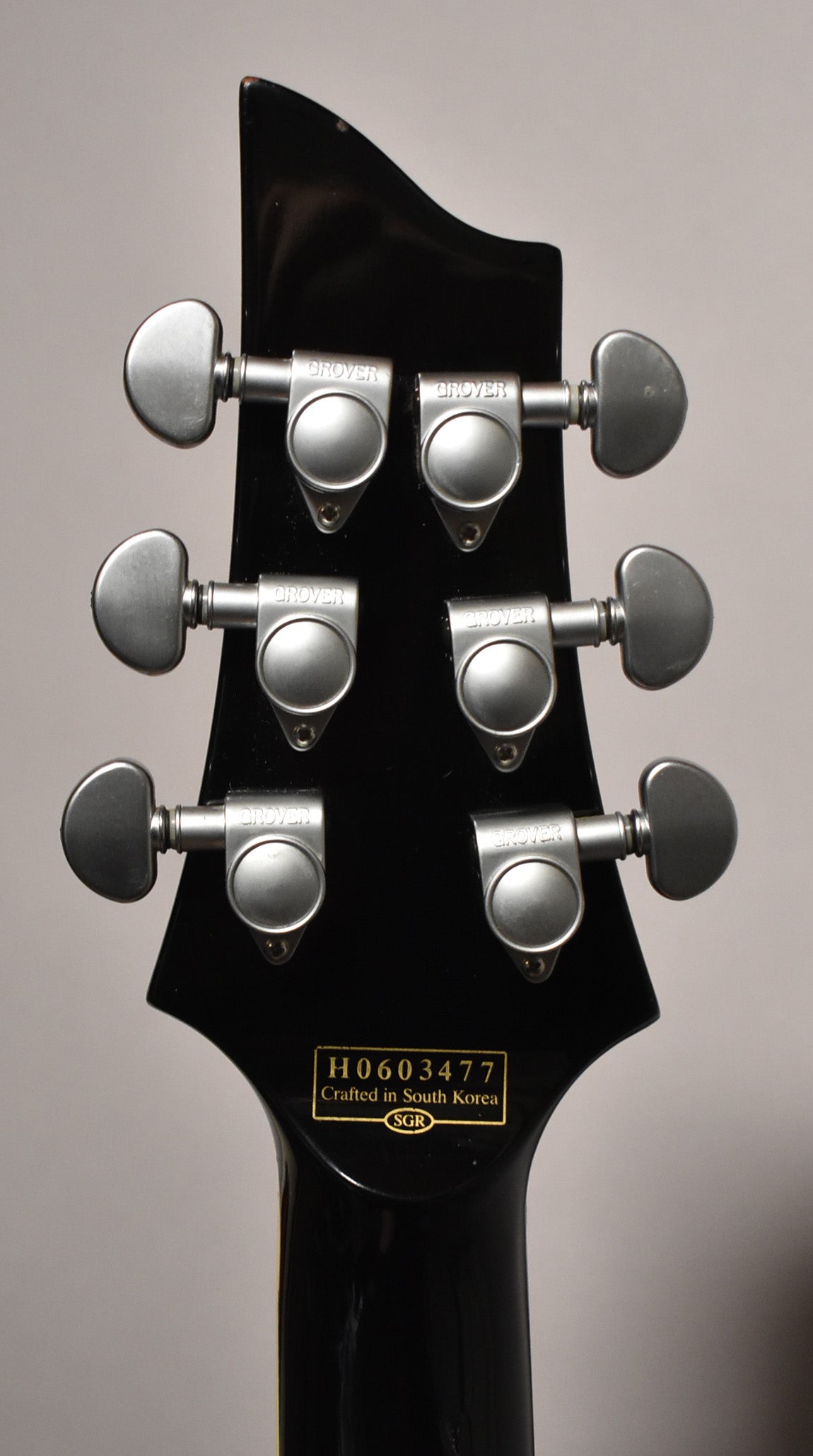 The back of a black gloss headstock with six chrome tuners, showing minor wear on the top edge.