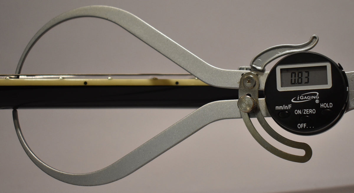 A digital caliper measures the guitar's black neck, showing a reading of 0.83, with the light-colored fretboard visible.