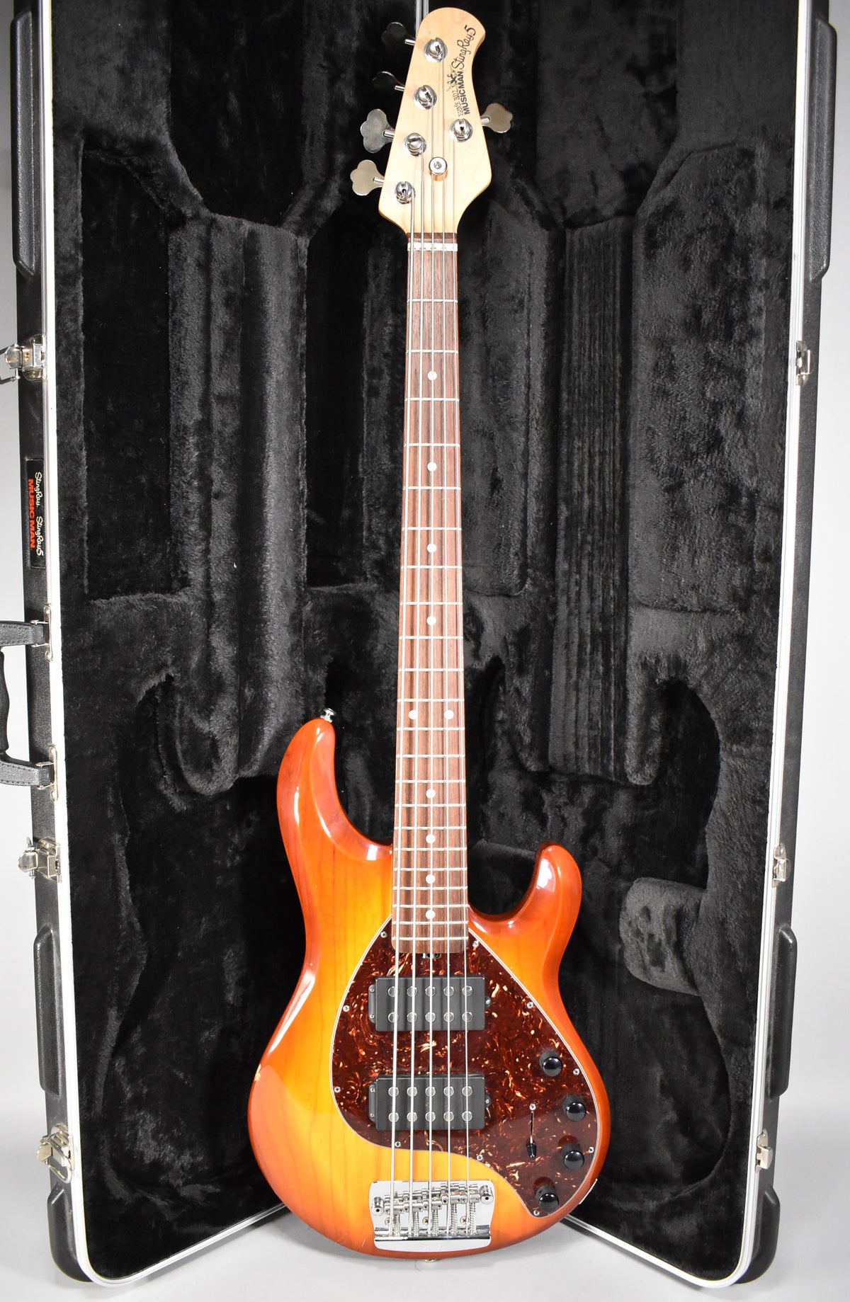 Full-length view of a honey sunburst 5-string bass with a tortoiseshell pickguard, dual humbuckers, and a maple neck, in a black hard case.