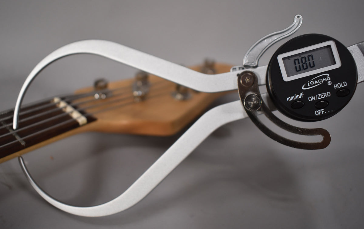 A digital caliper measures the bass's light brown headstock and fretboard from the side, showing the nut and tuning pegs.
