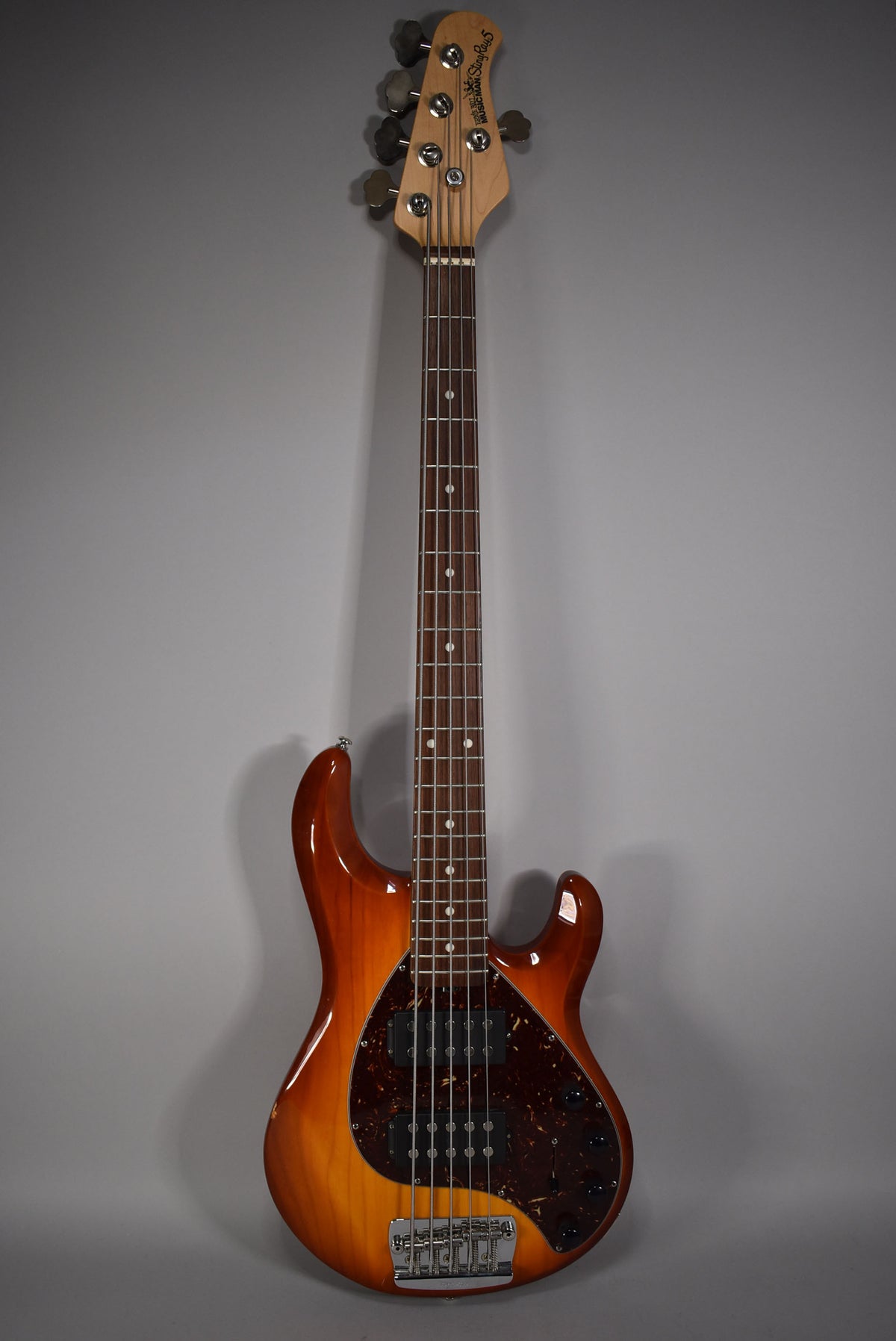 Full front view of a 5-string bass in honey sunburst, with a tortoiseshell pickguard, two humbucker pickups, and a chrome bridge.