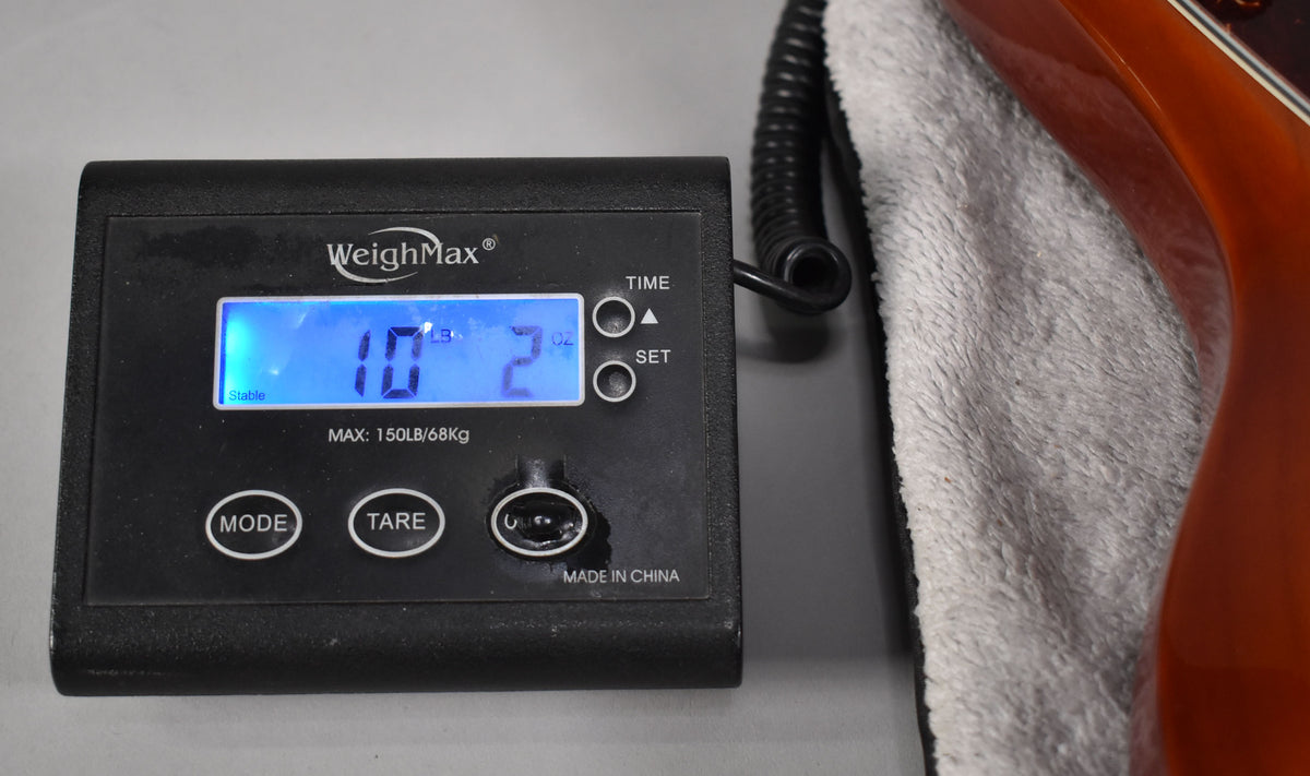 A digital scale displays "10 LB 2 OZ" next to the bass guitar's honey sunburst body, resting on a white towel.