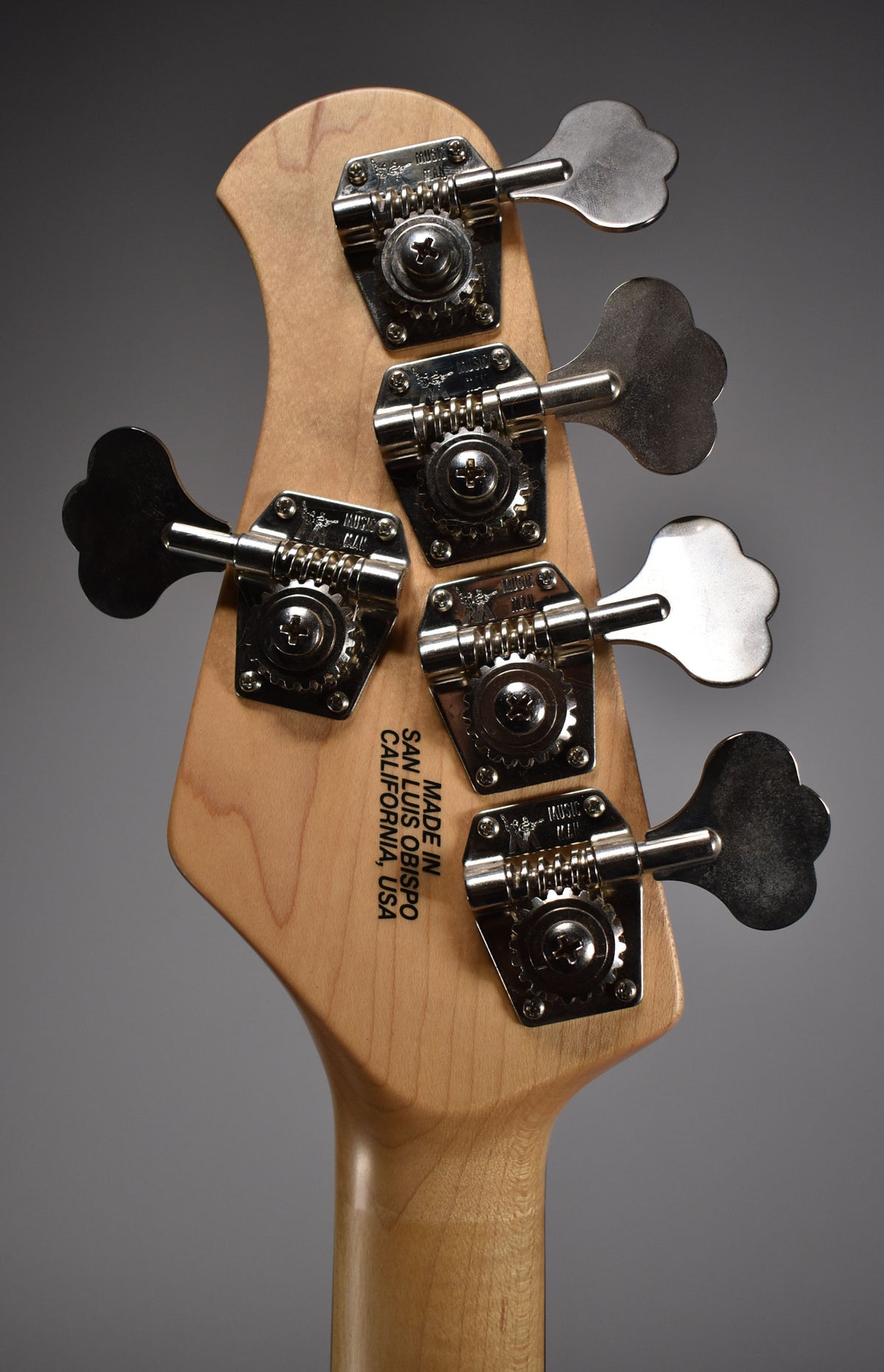 The back of a light wood bass guitar headstock with five chrome clover-leaf tuners, showing light wear.