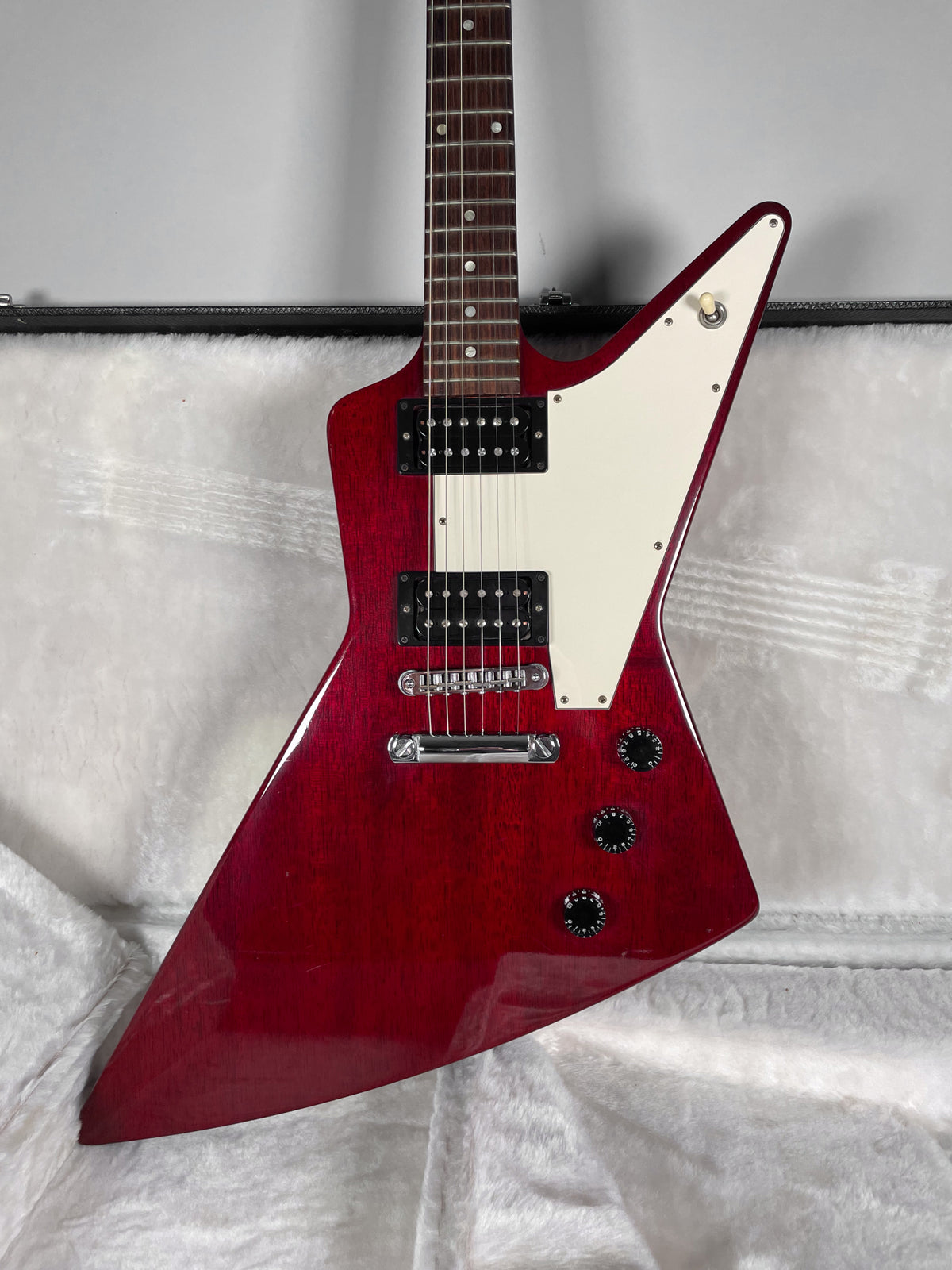 A wine red electric guitar with a white pickguard, two humbuckers, and three black control knobs, resting in a white plush case.