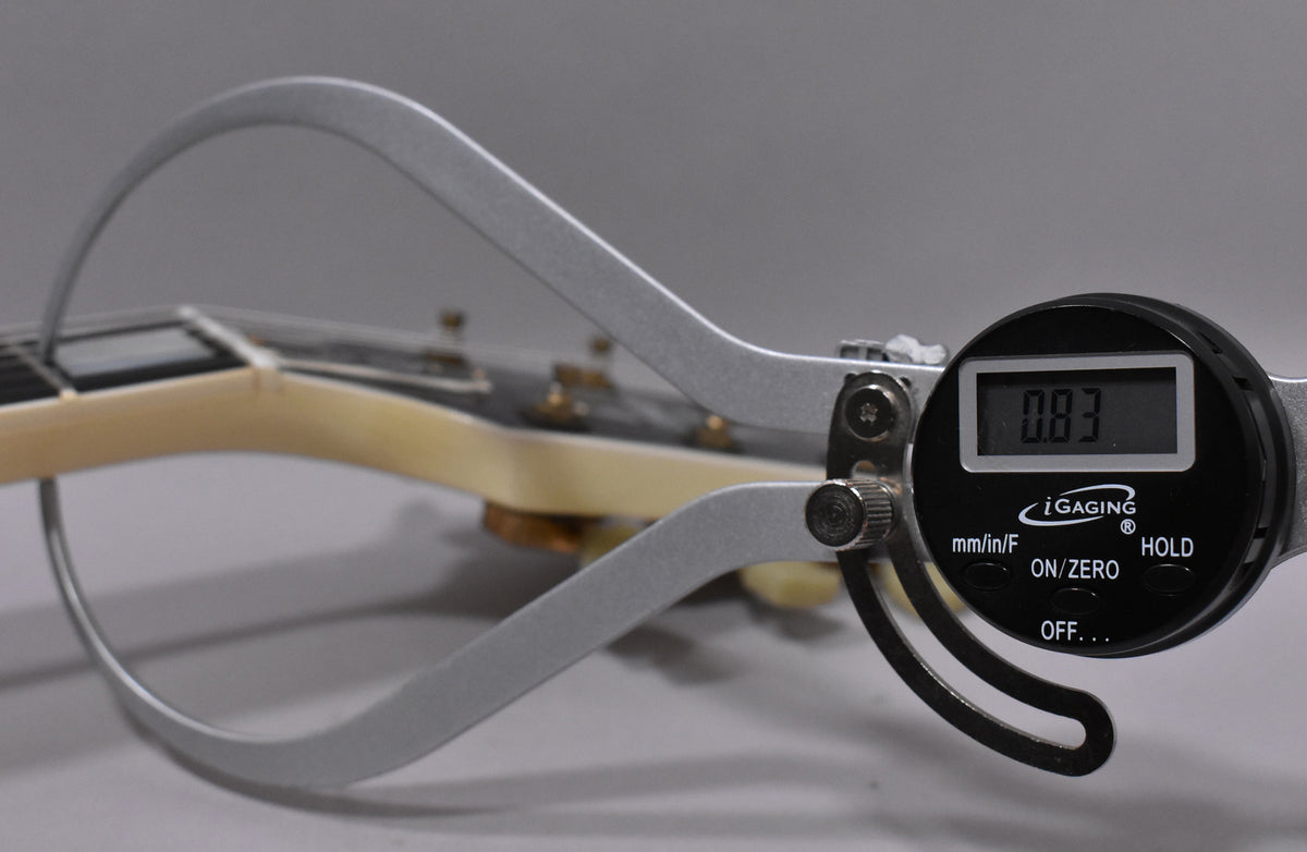 A digital caliper measures the guitar's nut width at 0.83 inches, with the headstock and tuning pegs blurred in the background.