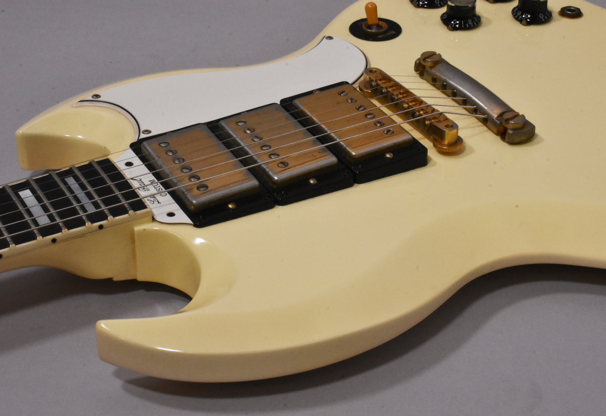 The cream SG body shows three gold-covered humbuckers, a white pickguard, and a gold bridge with a separate tailpiece.