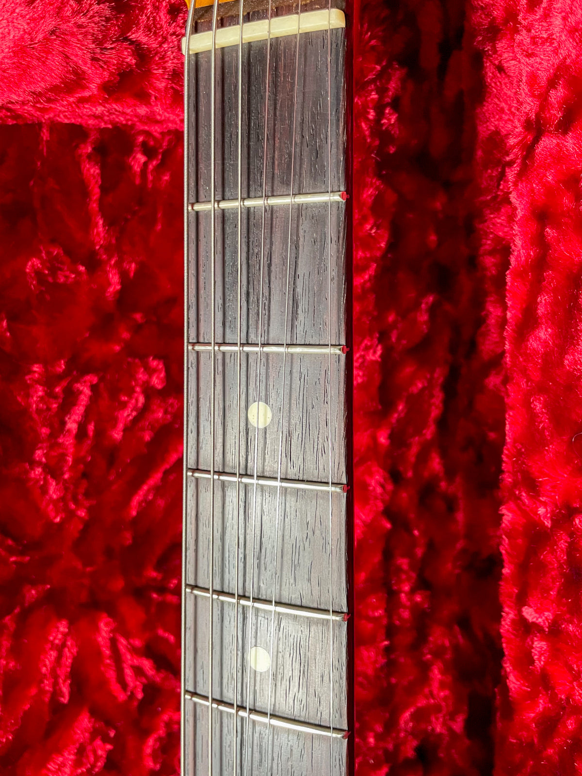 Close-up of the dark fretboard with light dot inlays and frets, angled against a plush red case interior.