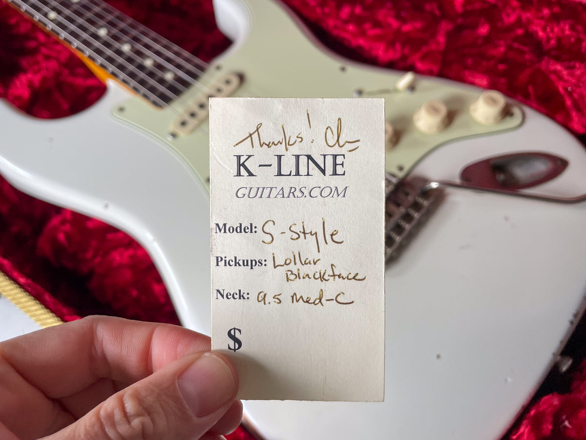 A hand holds a white tag over an Olympic White relic guitar with a mint green pickguard and aged white controls, resting in a red velvet case.