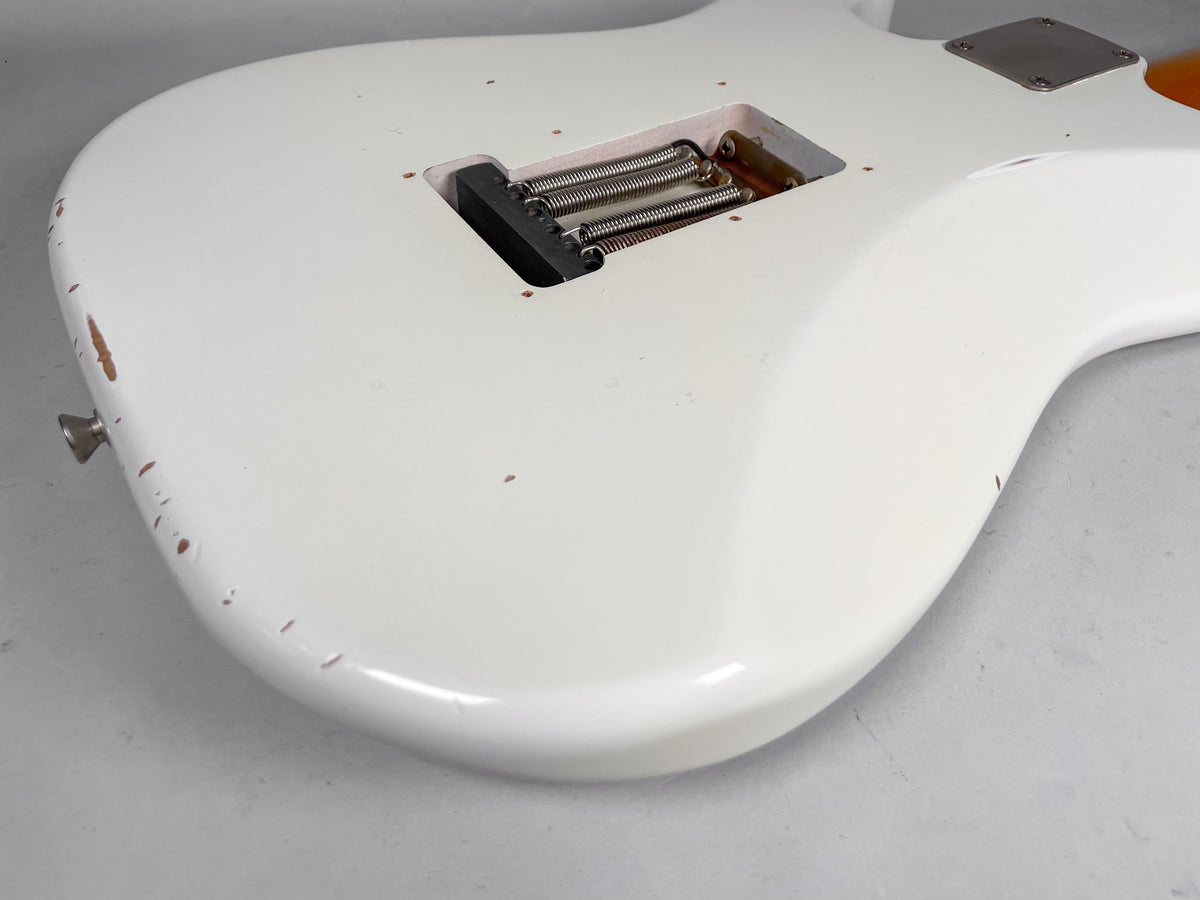 The back of an Olympic white Strat-style guitar shows a tremolo spring cavity, strap button, and heavy relic wear with paint chips.