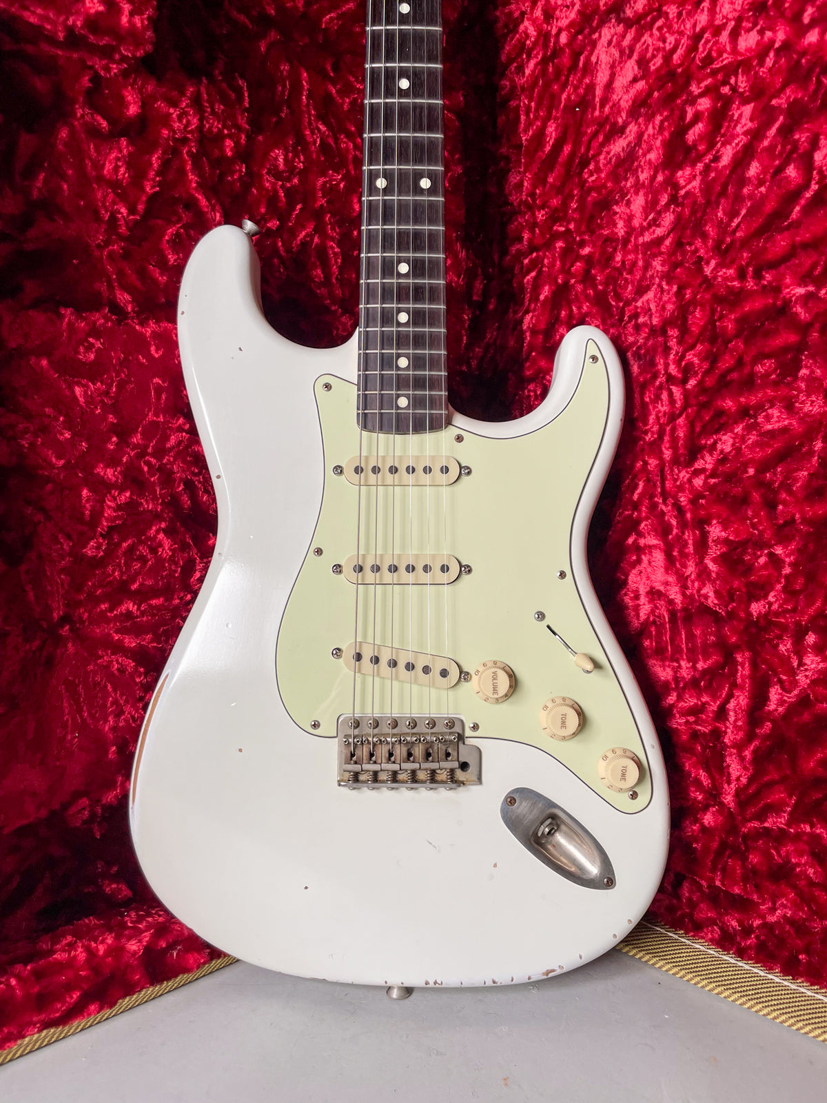 An Olympic White Strat-style guitar with a mint green pickguard and aged cream hardware, showing relic wear, on red velvet.