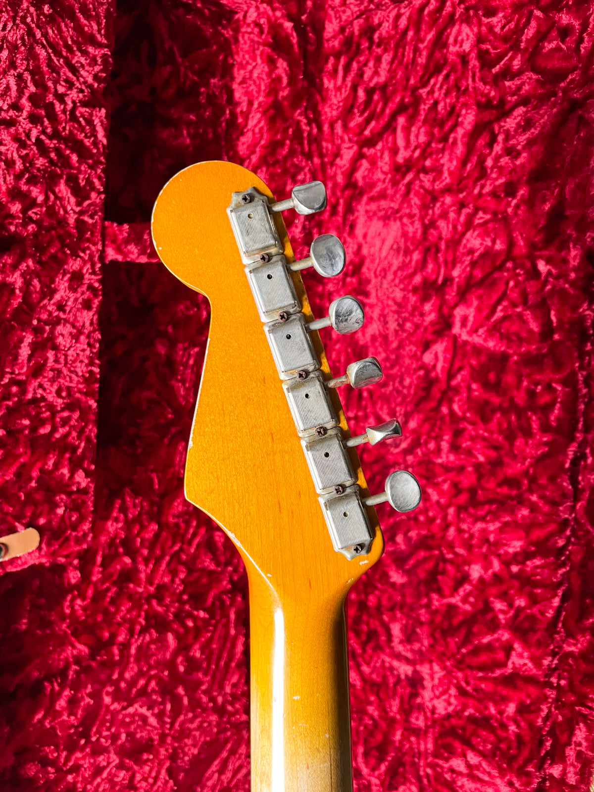 The back of the guitar's amber-tinted headstock and neck, showing six worn, rectangular tuners, against a red velvet case lining.