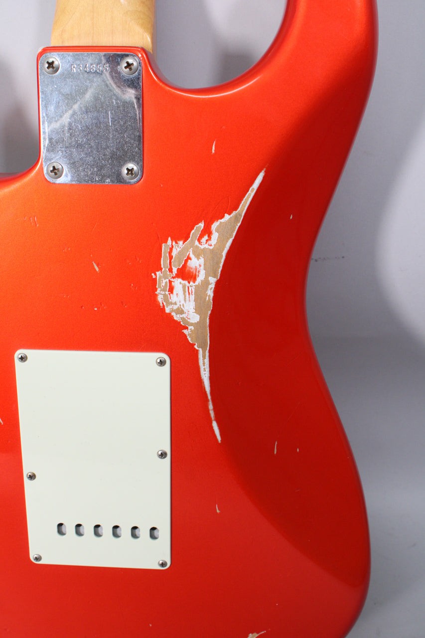 The back of a metallic orange Stratocaster body, showing a large area of chipped paint exposing wood, and a white tremolo cover.