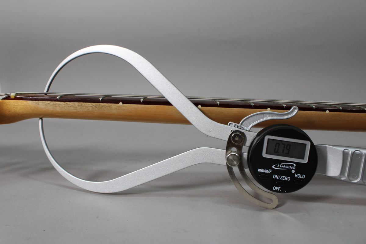 A digital caliper measures the guitar's neck thickness at 0.79, showing the light wood neck and dark fretboard with dot inlays.