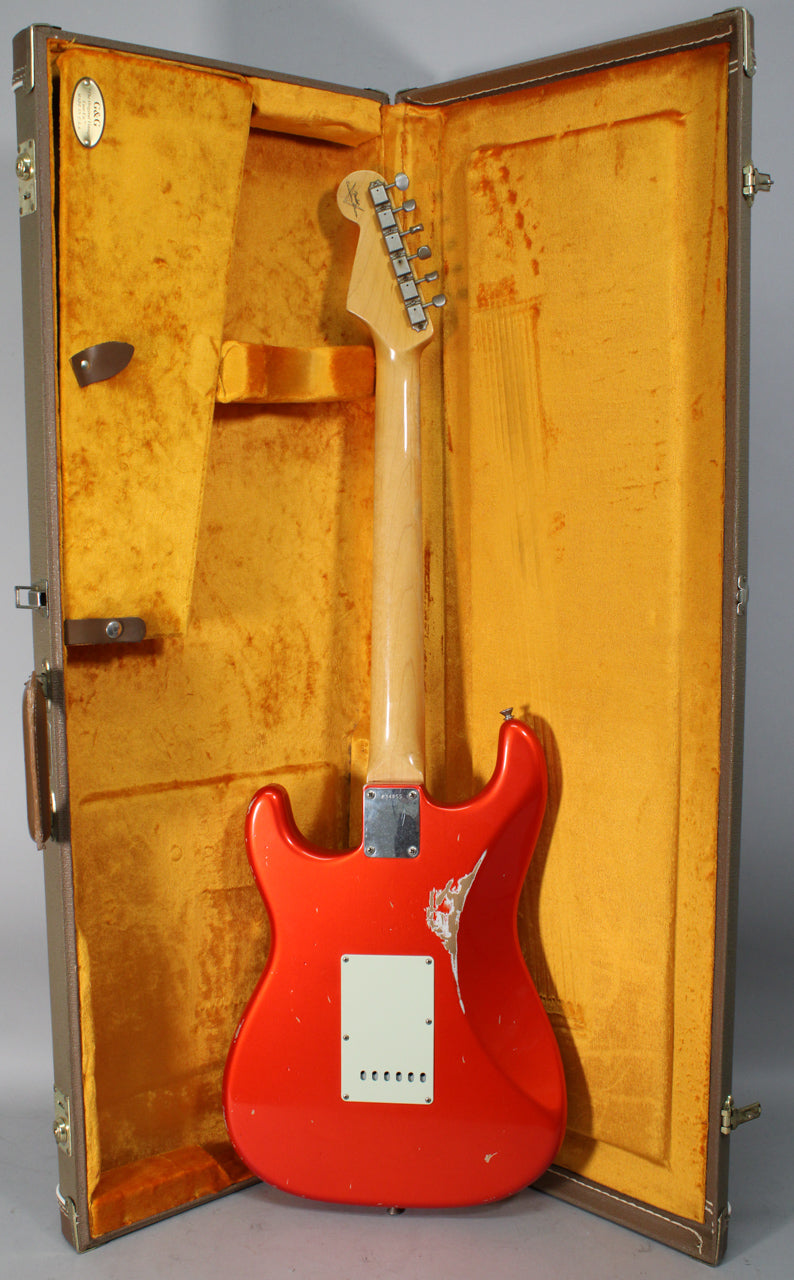 The back of a bright orange Stratocaster with a relic finish, showing a large chip near the lower horn, in a brown case with gold lining.