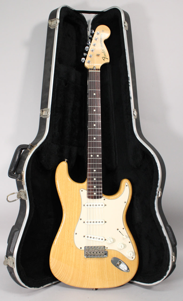 A full-front view of a natural-finish Stratocaster with a white pickguard, rosewood fingerboard, and chrome hardware, resting in its open black hard case.