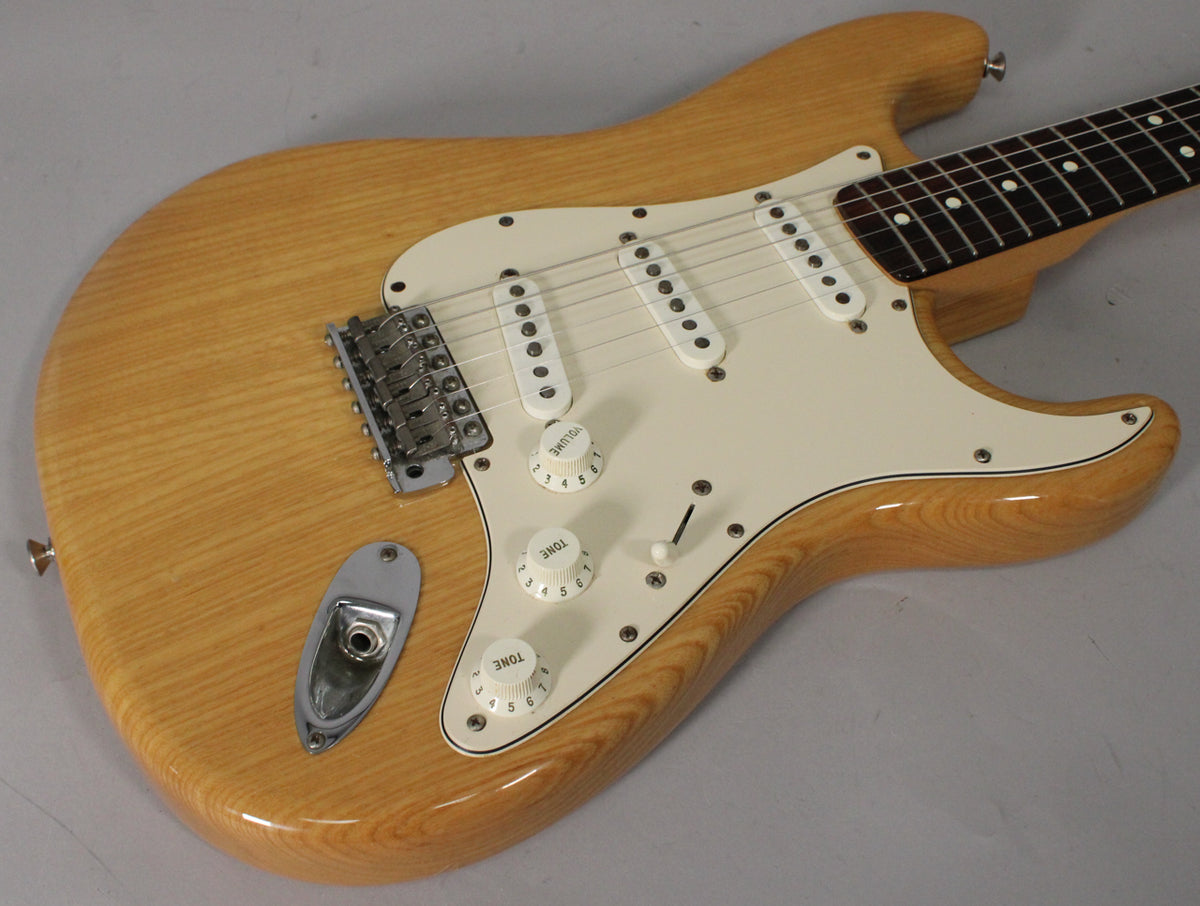 The natural wood grain body of the Stratocaster is visible from the upper bout to the lower horn, with a white pickguard and three single-coil pickups.