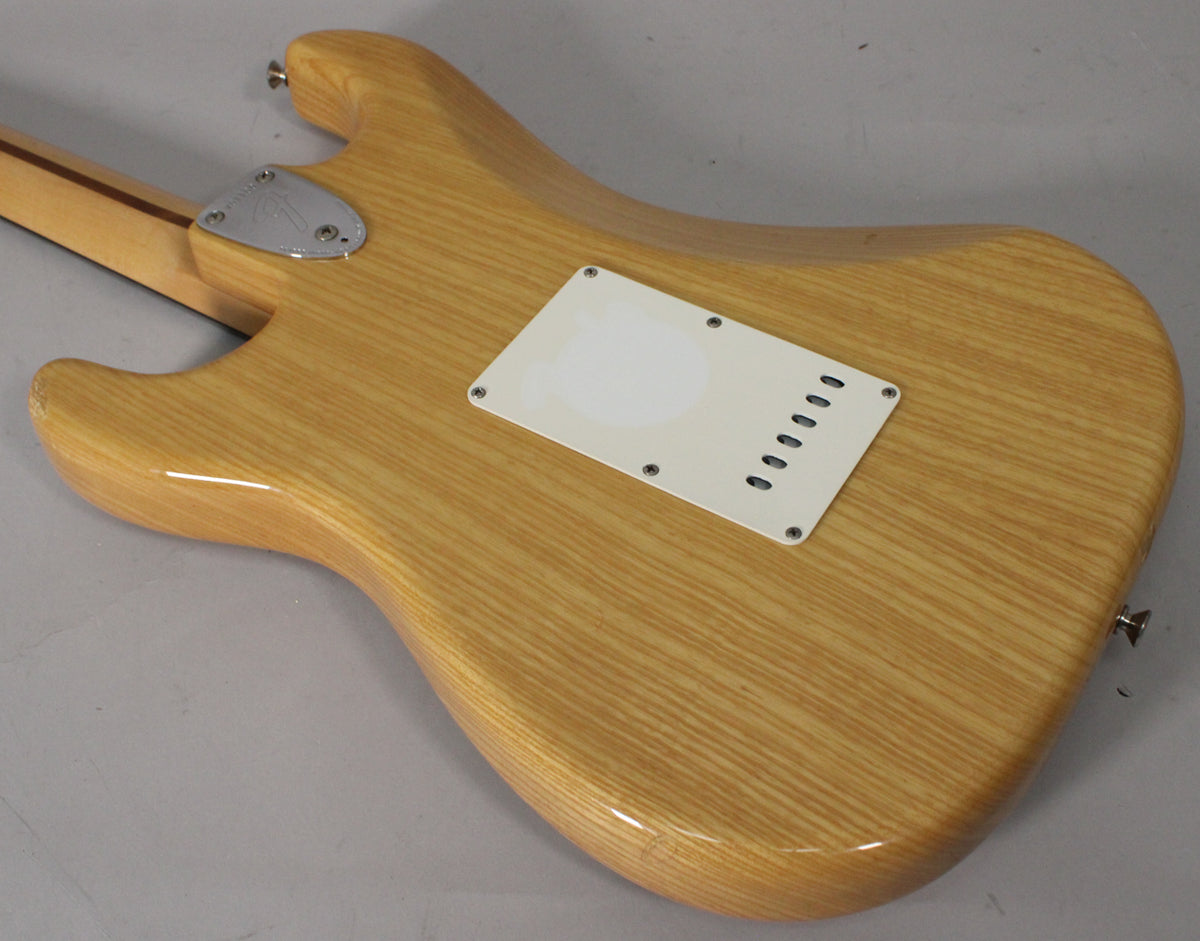 The back of a light blonde Stratocaster body with visible wood grain, a white tremolo cover, and a chrome neck plate.