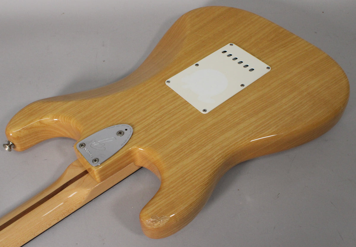 The back of a light blonde Stratocaster body with a white tremolo cover, a chrome neck plate, and a chip on the lower horn.