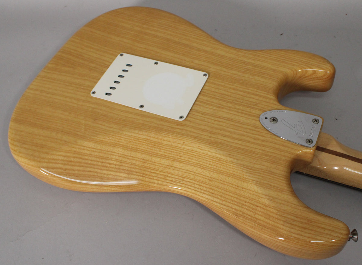 The back of a light wood Stratocaster body with a white tremolo cover and chrome neck plate, showing light wear.