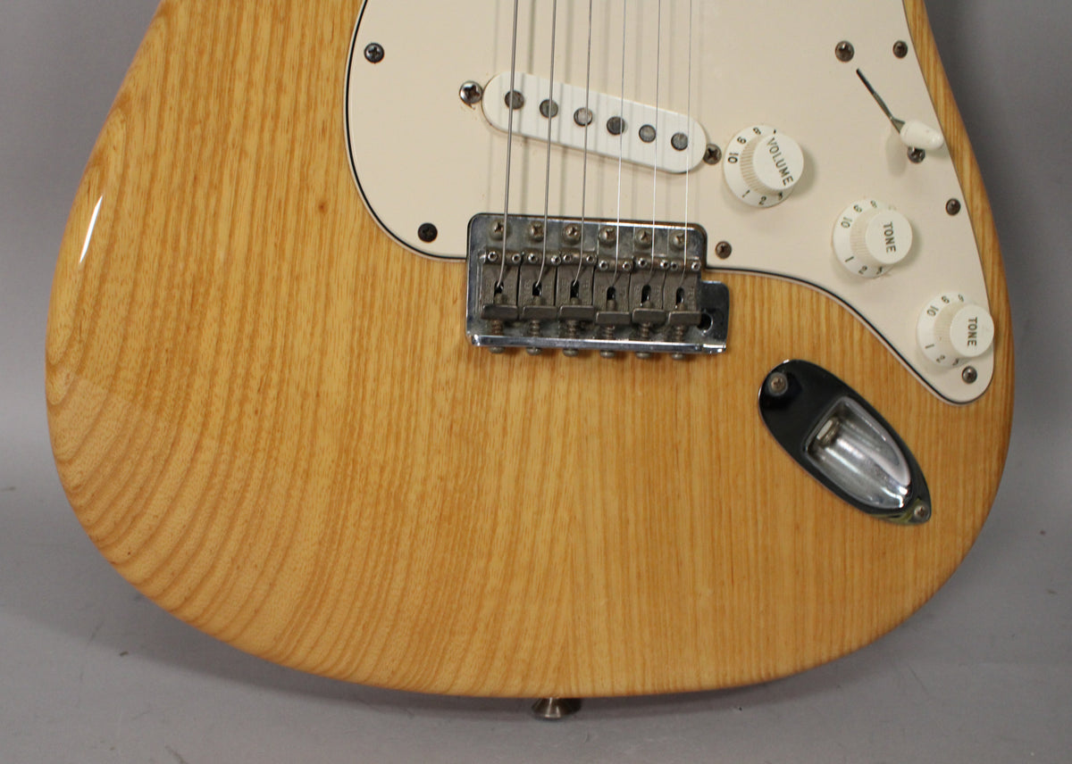 Close-up of a natural ash Stratocaster body with a cream pickguard, chrome bridge, white pickups, and control knobs.