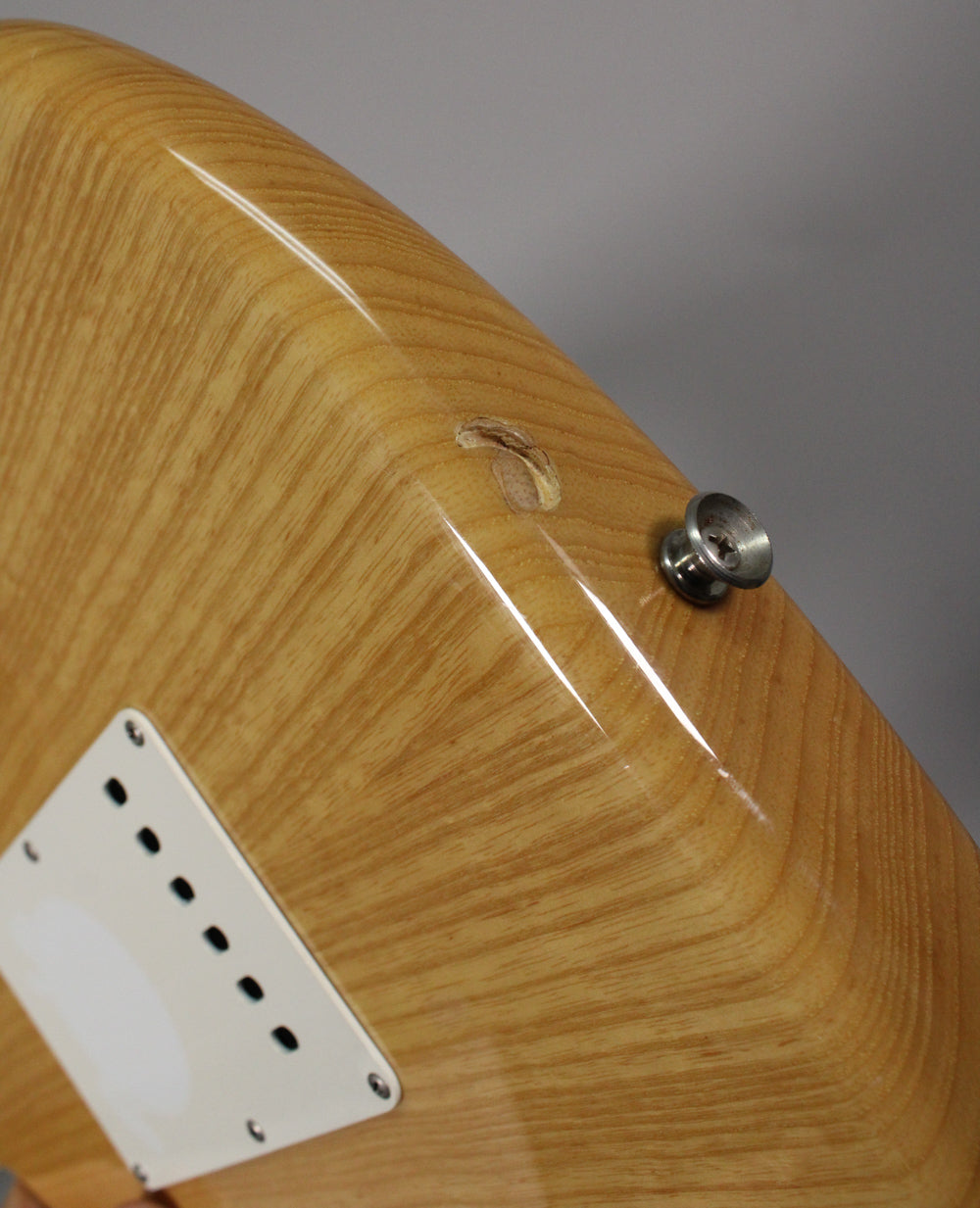 Close-up of the guitar's natural wood grain body, showing a white tremolo cover, a strap button, and a chip in the wood near the button.