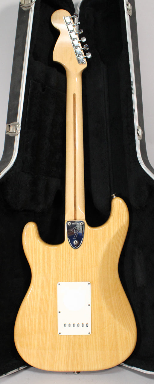 The back of a natural finish Stratocaster with a maple neck, chrome tuners, and a white tremolo cover, in a black hard case.