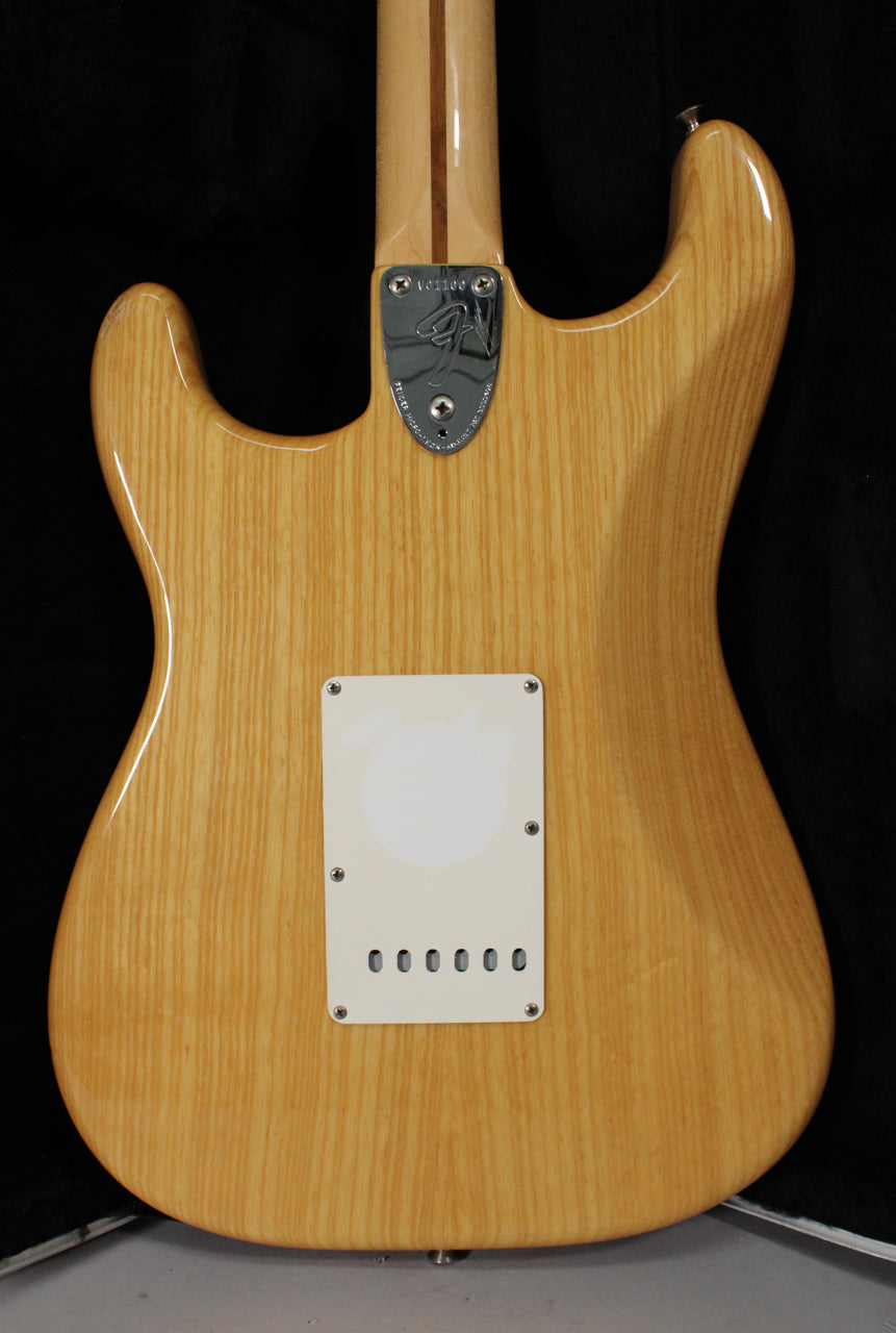 Rear view of a natural finish Stratocaster with visible wood grain, a white tremolo cover, and a chrome neck plate.