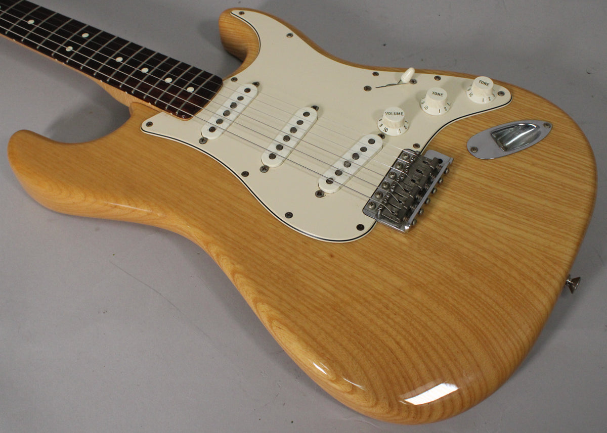 The natural finish Stratocaster body and neck are angled, showing the cream pickguard, three single-coil pickups, and chrome tremolo bridge.