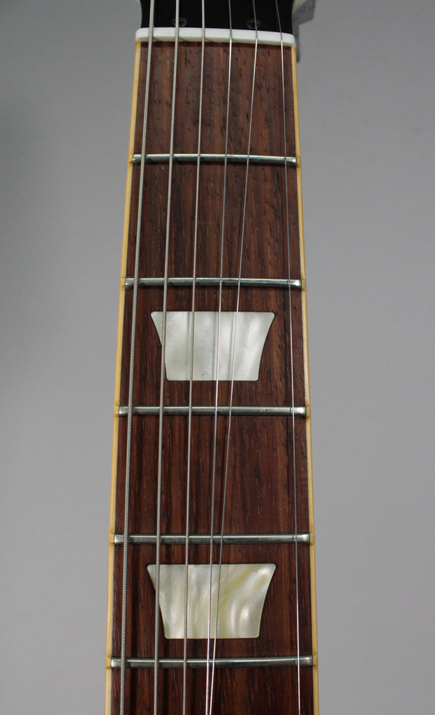 Close-up of the dark brown fretboard with two trapezoidal mother-of-pearl inlays, silver frets, and six strings.