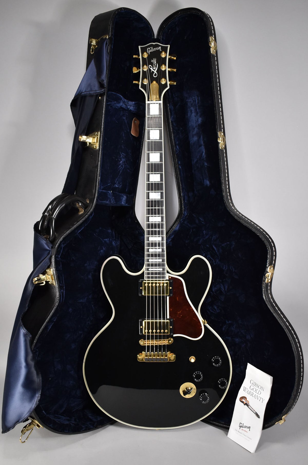A full-length, glossy black electric guitar with gold hardware and a dark tortoiseshell pickguard, resting in a blue velvet-lined hard case.