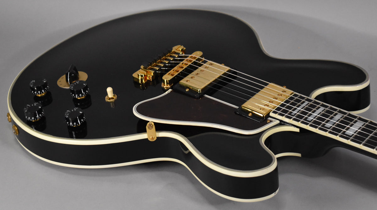 The upper body of a glossy black electric guitar with gold hardware, a dark pickguard, and white binding.