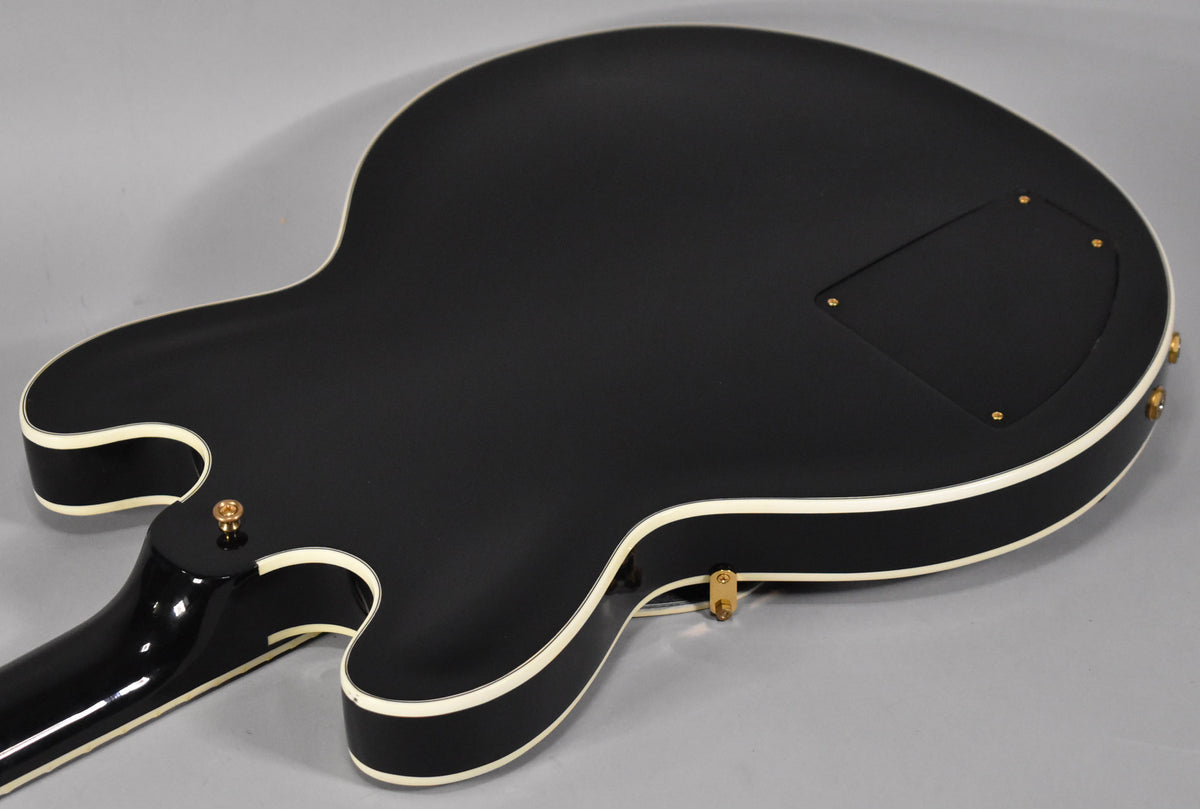 The back of an ebony semi-hollow guitar with white binding, showing a control cavity cover and gold strap buttons.
