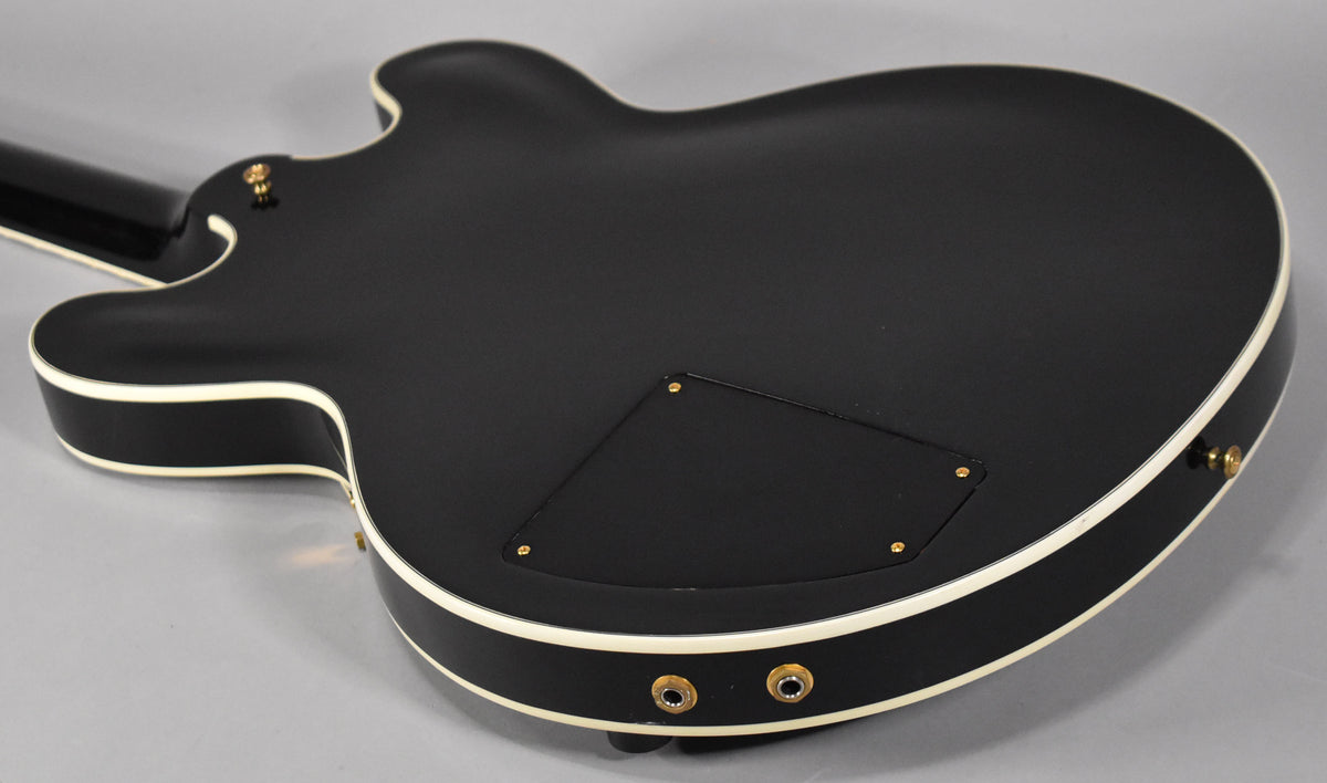 The back of an ebony guitar with a matte finish and cream binding, showing the control cavity cover and two output jacks.