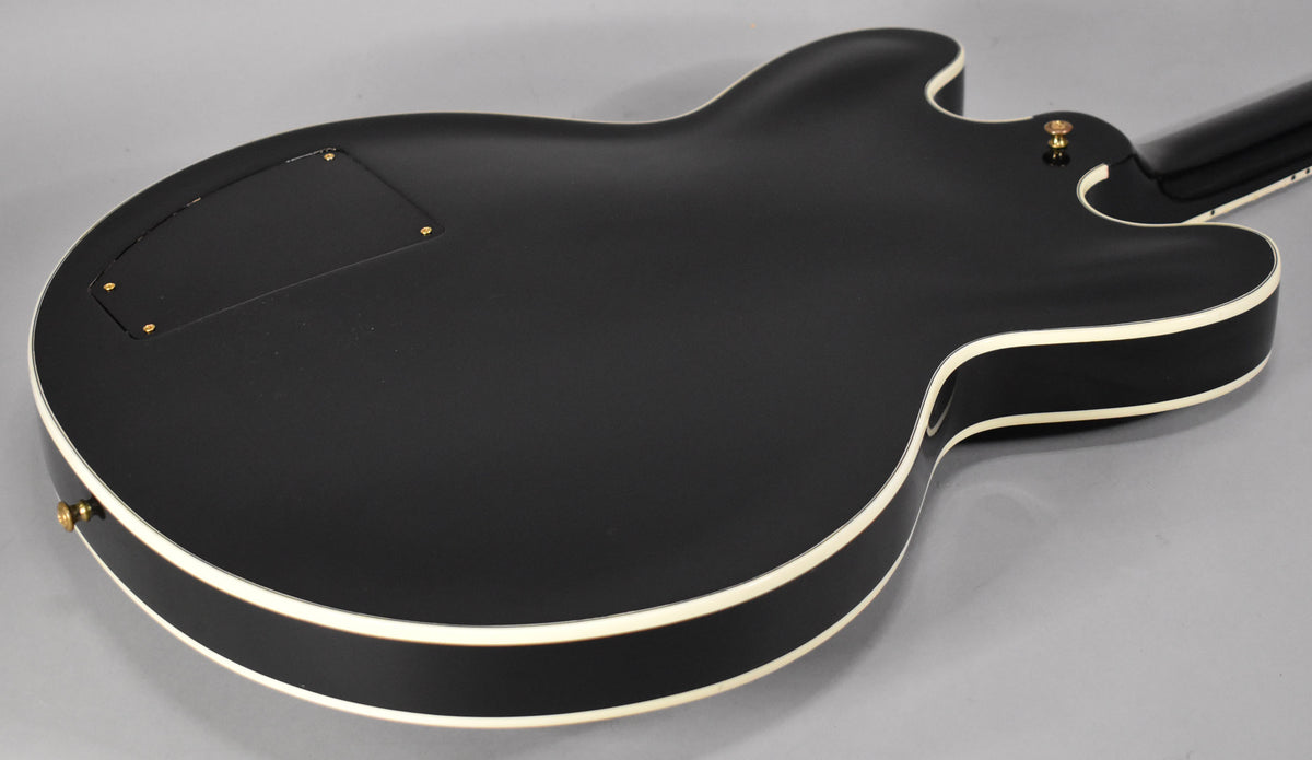 The back of a black semi-hollow guitar with white binding, showing a control cavity cover and a strap button.