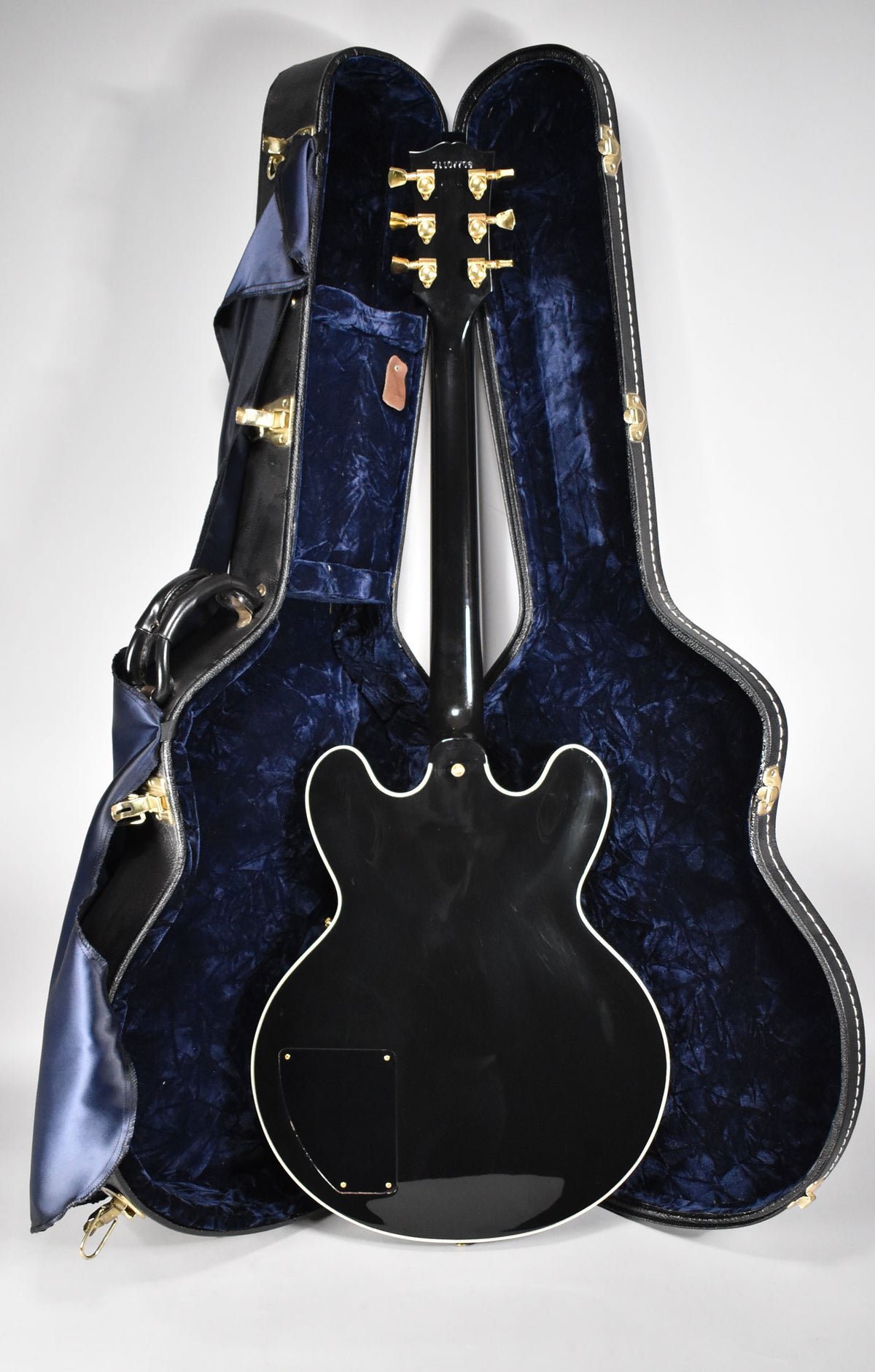 The ebony guitar's back is visible, with a black control plate, white binding, and gold tuners, resting in a blue velvet-lined case.
