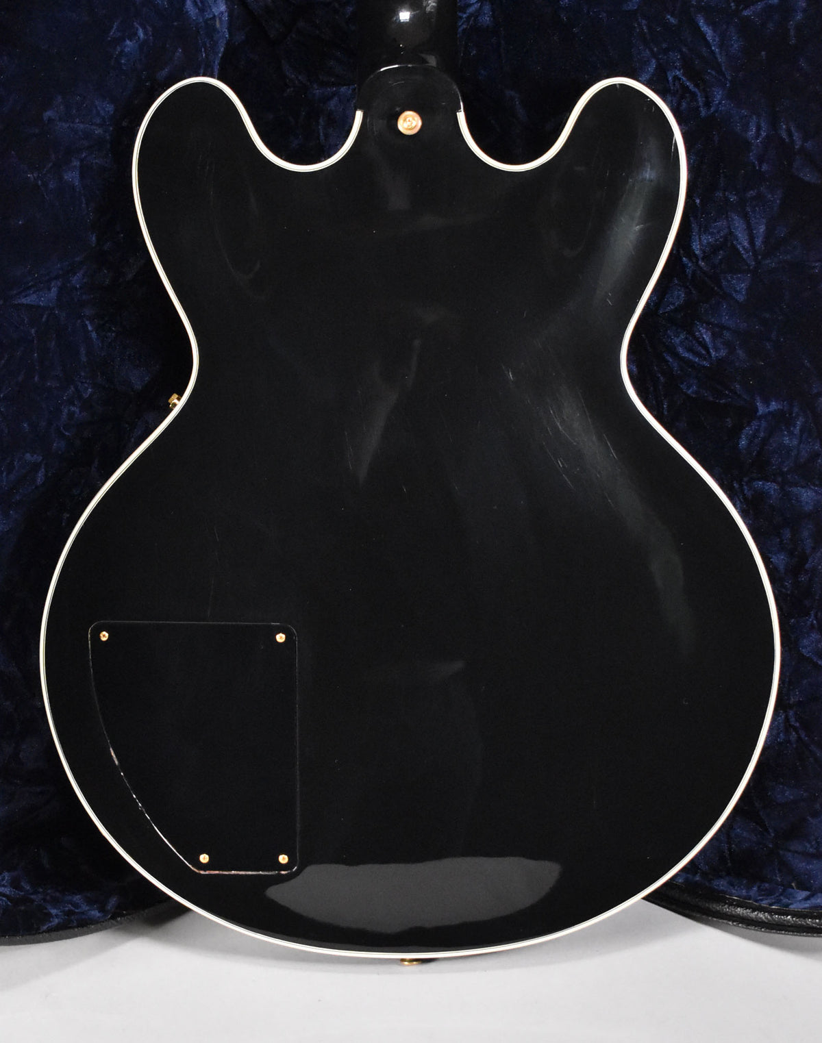 The back of a glossy black hollow-body guitar with white binding, showing light scratches and a gold-screwed control plate.