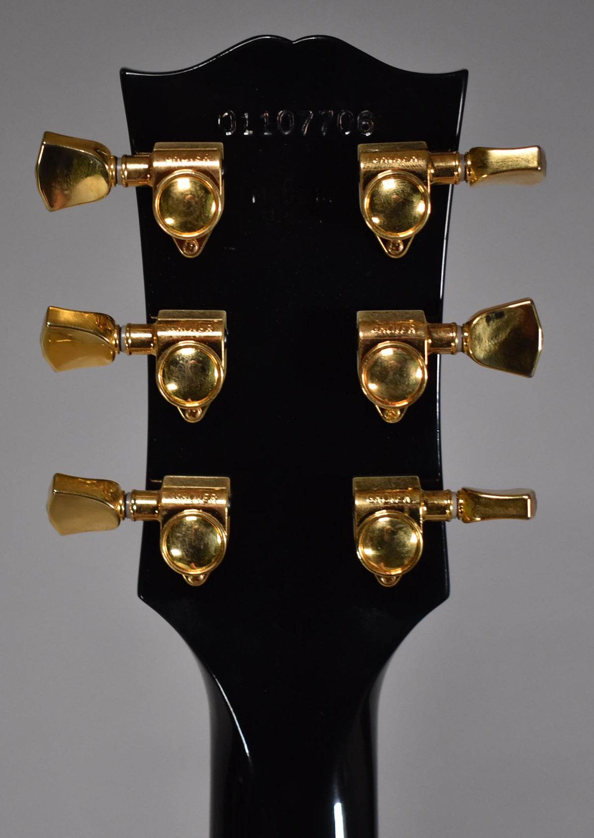 The back of an ebony headstock with six gold Grover tuners, showing light wear and a faint serial number.