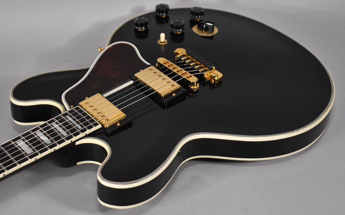 Close-up of a glossy black hollow-body guitar with gold hardware, showing the lower bout, pickups, bridge, and part of the fretboard.