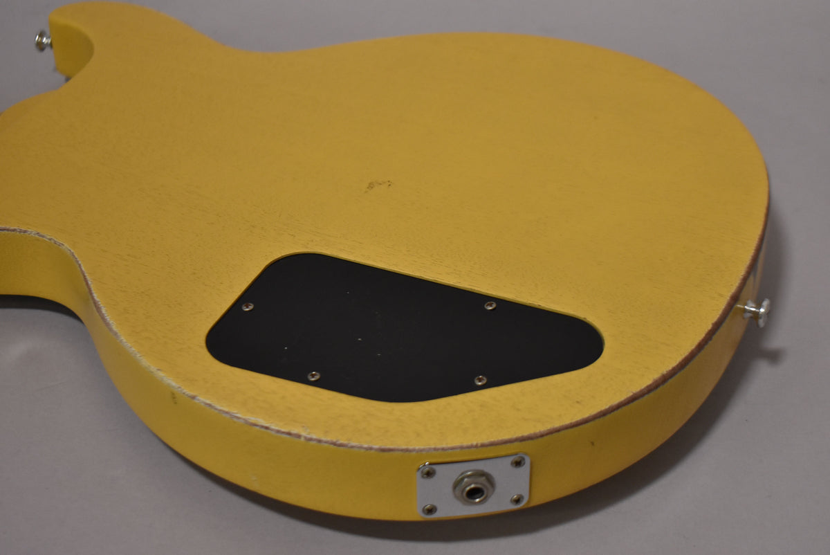 The back of a faded TV yellow guitar body, showing a black control cavity cover and a chrome output jack plate with wear on the edges.