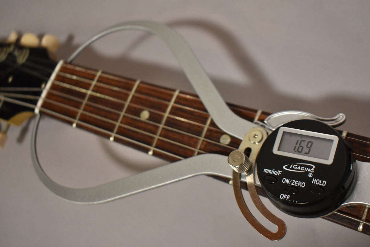 A digital caliper measures the nut width of the dark brown fretboard, showing 1.69 inches on its display.