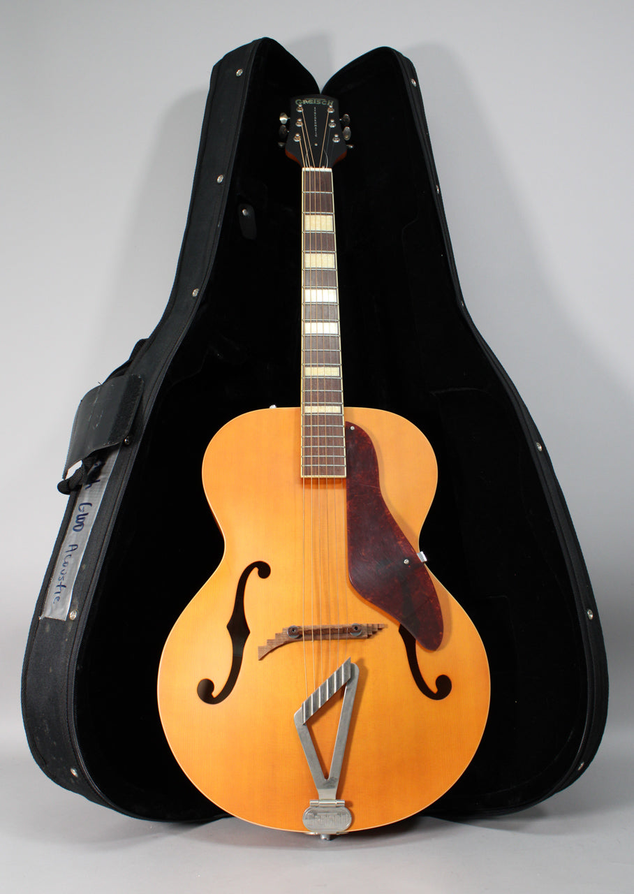 Full-length view of a natural matte archtop guitar with f-holes, a dark pickguard, and a chrome trapeze tailpiece, in its open case.