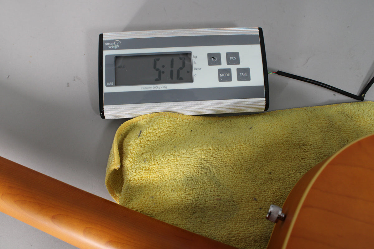 A digital scale displays "5.12" above a yellow cloth, with the guitar's natural matte neck and body visible below.