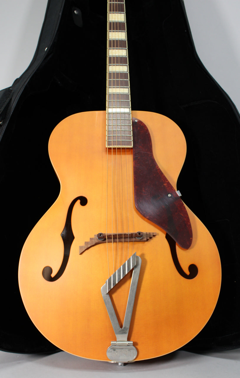 Full front view of a natural matte archtop guitar, showing the F-holes, tortoise pickguard, and trapezoid tailpiece.